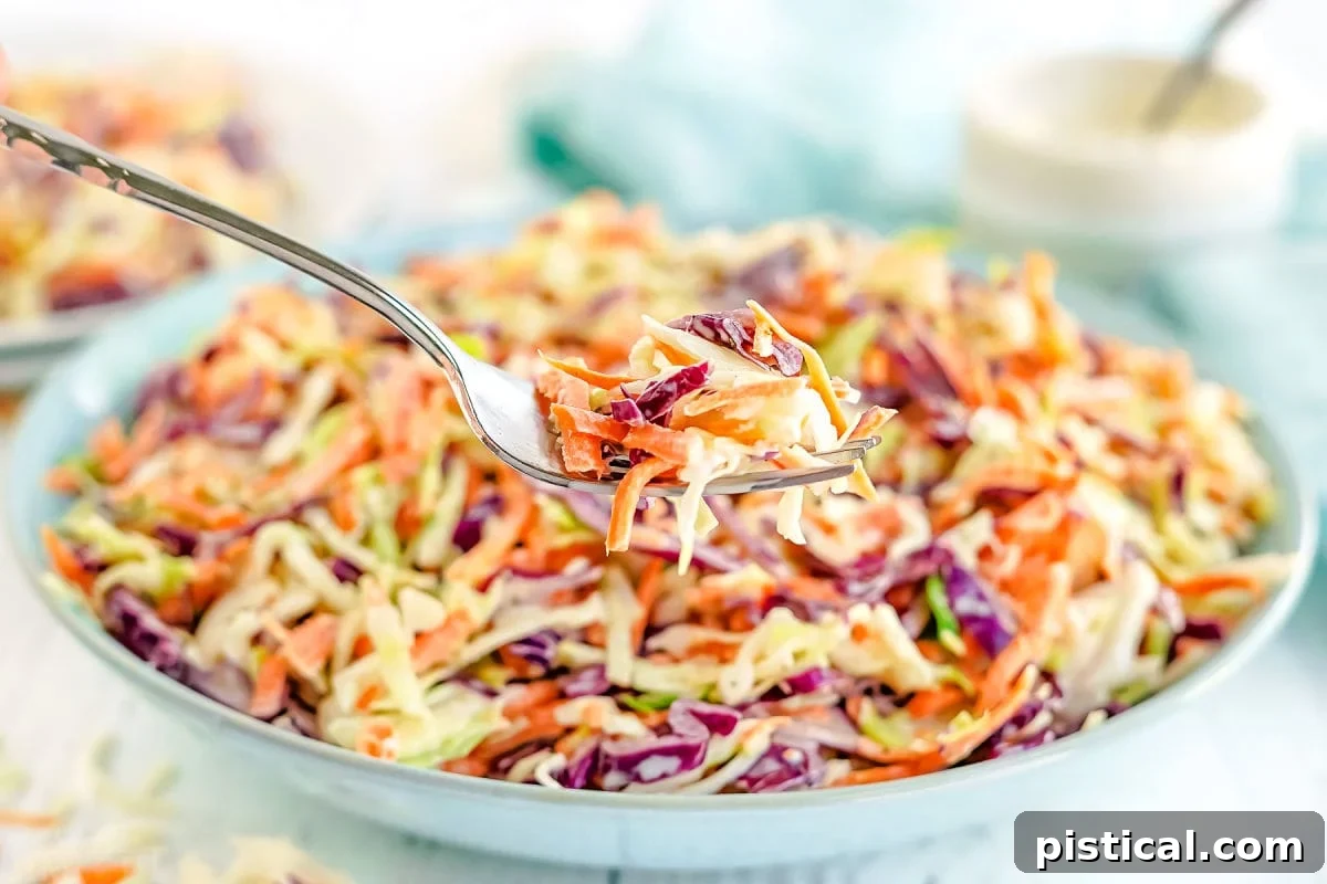 A fork holding a generous serving of creamy coleslaw, with a large bowl of coleslaw visible in the soft-focused background.