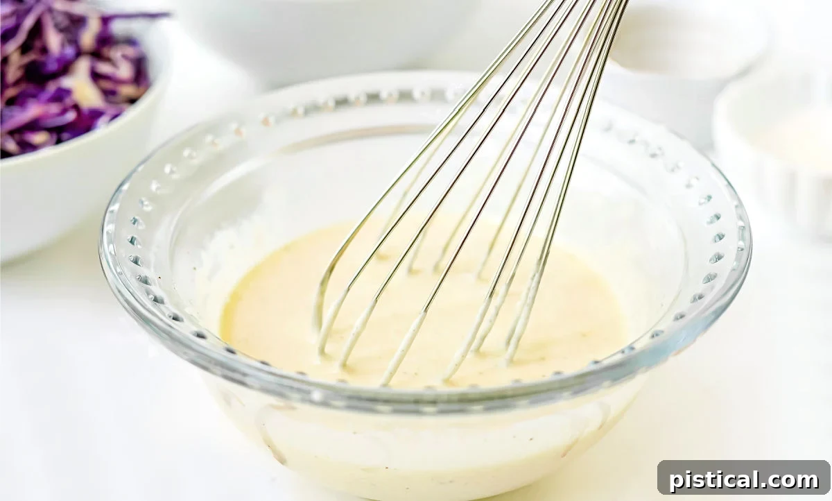 A glass bowl filled with creamy coleslaw dressing, accompanied by a whisk, ready to be mixed with fresh vegetables.