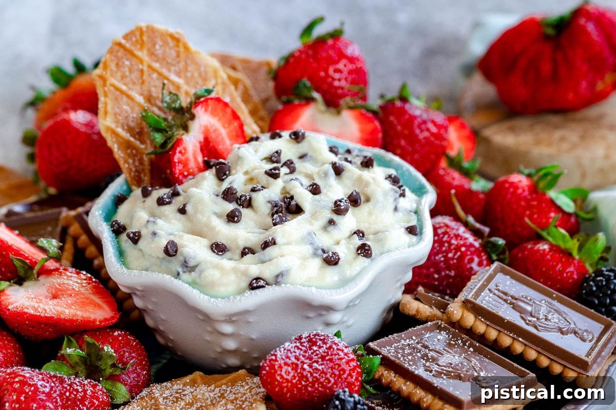 Decadent Cannoli Dip 3 A small white bowl with a charming blue interior holds creamy Cannoli Dip generously sprinkled with mini chocolate chips, surrounded by an assortment of golden cookies and fresh, vibrant strawberries, all poised for dipping pleasure.