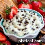 Creamy Cannoli Dip, generously garnished with mini chocolate chips, elegantly presented in a small white bowl with a charming blue interior, surrounded by a delightful selection of golden cookies and fresh, vibrant strawberries, all ready to be savored.