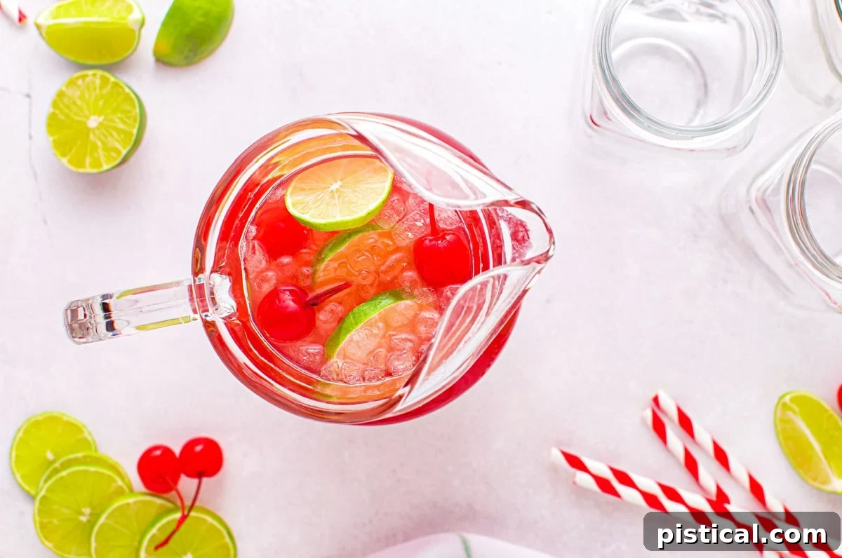 A top-down photographic perspective capturing various ingredients being gracefully combined and mixed into a large, clear glass pitcher to prepare the cherry limeade.