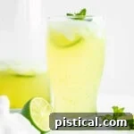 glass of limeade sitting in front of tall glass pitcher with more limeade in it. Fresh lime slices and mint sprigs scattered about.