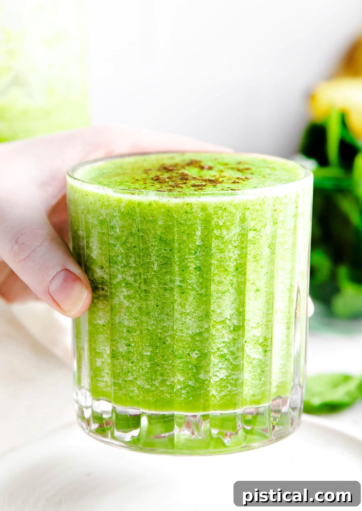 A hand holding a vibrant green smoothie in a ridged glass, with lush green foliage blurred in the background, emphasizing freshness.