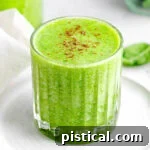 A simple green smoothie recipe in a glass, resting on a small white plate in front of a blender, emphasizing the freshness and simplicity.