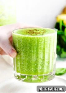 Hand holding a ridged glass filled with a refreshing green smoothie, against a blurred green background.