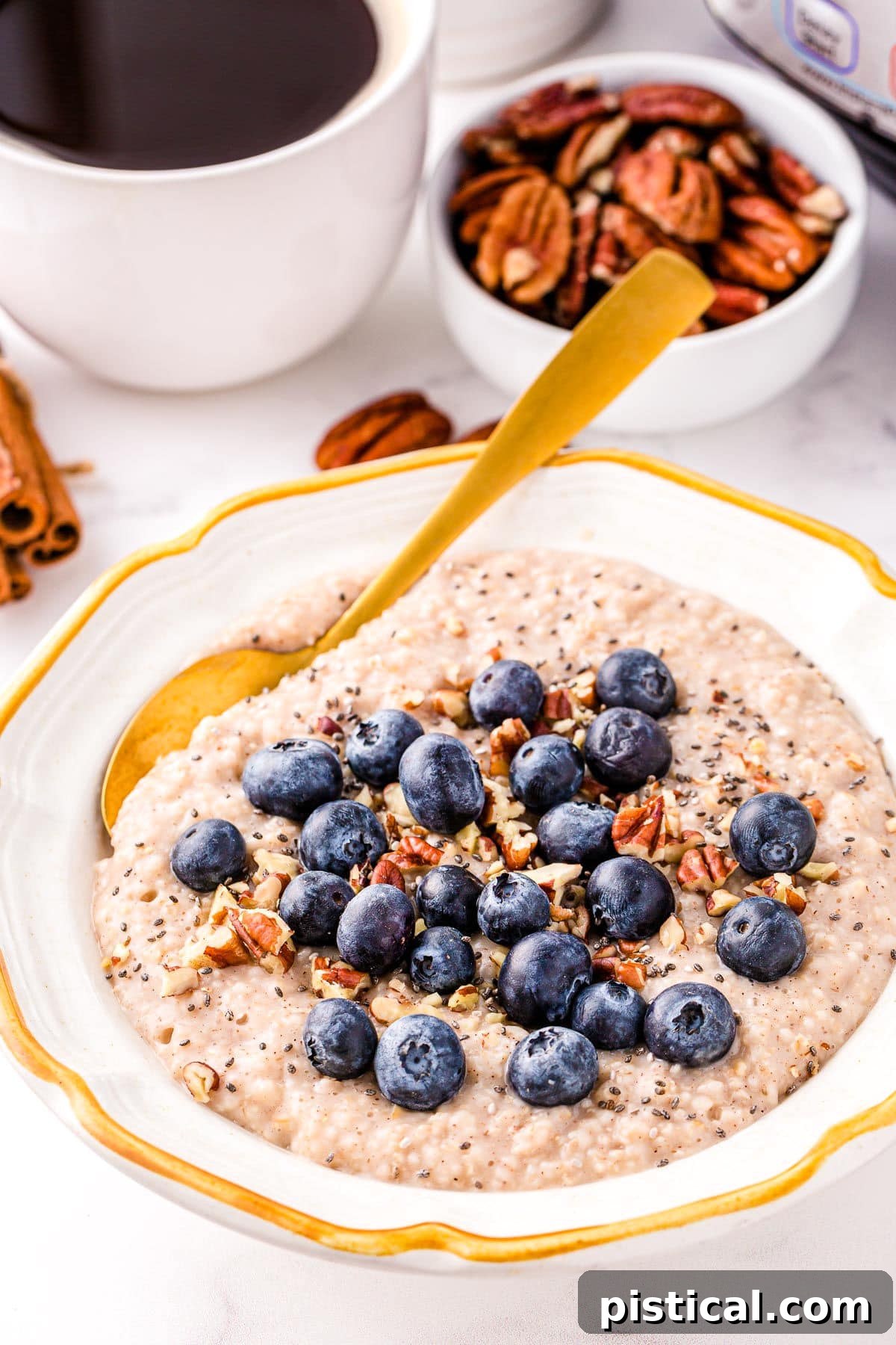 Instant Pot oats in a white bowl with a gold rim topped with blueberries and ground cinnamon. A cup of coffee and a small bowl of pecans are in the background, creating a cozy breakfast scene.