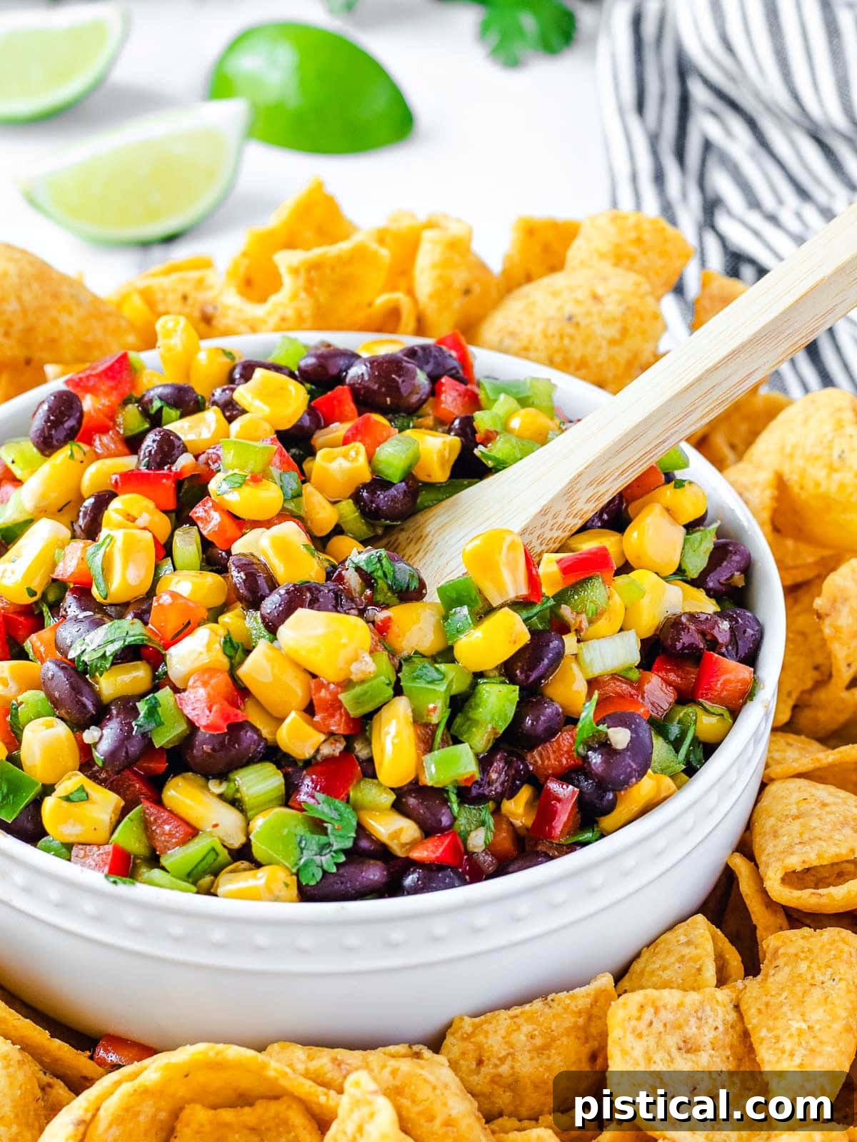 A large white bowl filled with colorful black bean and corn salsa, featuring vibrant red and green bell peppers, golden corn, and dark black beans. A wooden spoon rests in the salsa, surrounded by golden tortilla chips and fresh lime wedges in the background.