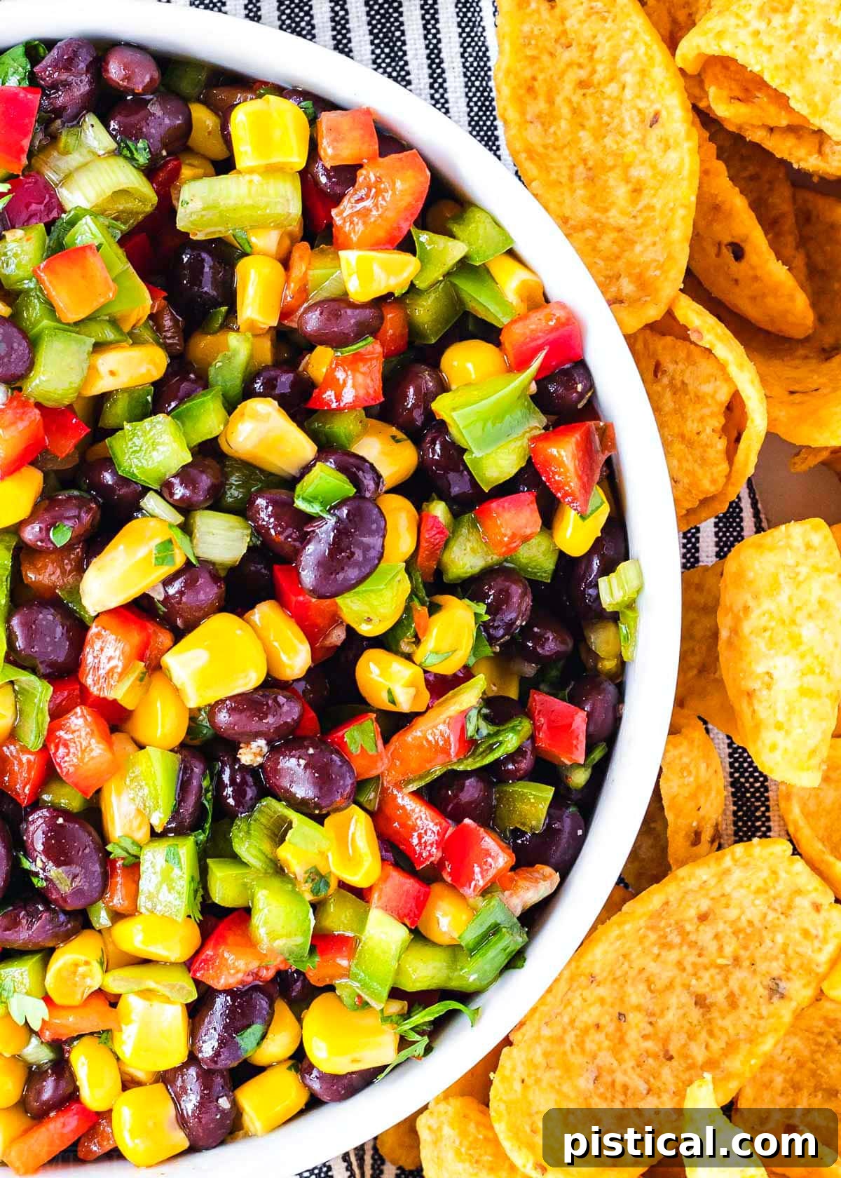 A close-up view of the vibrant and colorful black bean and corn salsa in a white serving bowl. The rich hues of red bell pepper, green bell pepper, yellow corn, and black beans are clearly visible, with tortilla chips scattered around the bowl for serving.