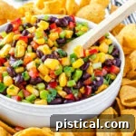 corn salsa with black beans in white bowl and wood spoon inserted. corn chips surrounding the bowl with limes in the background.