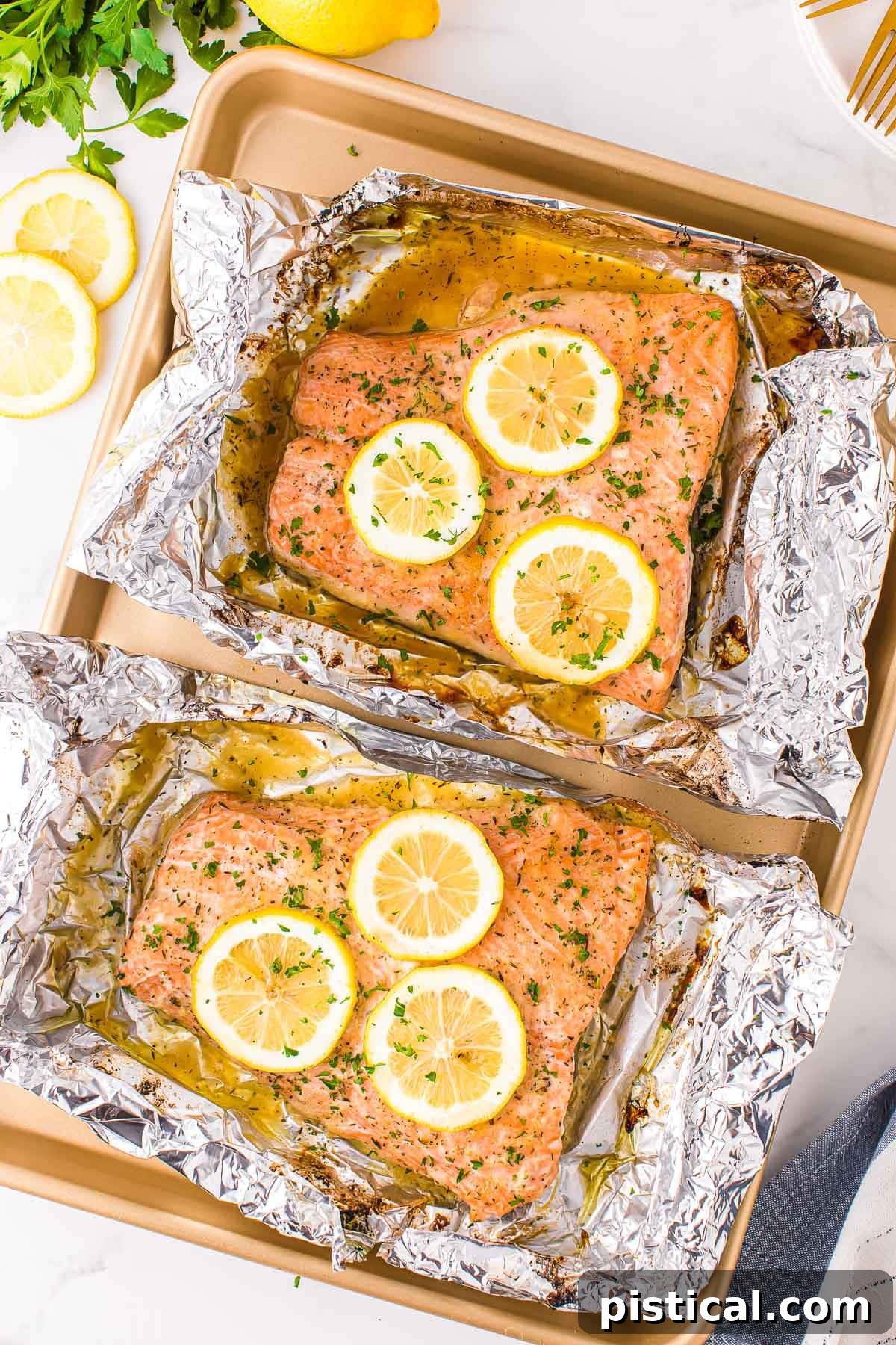 two large salmon filets baked in foil sitting on gold sheet pan topped with lemon slices and fresh minced parsley.
