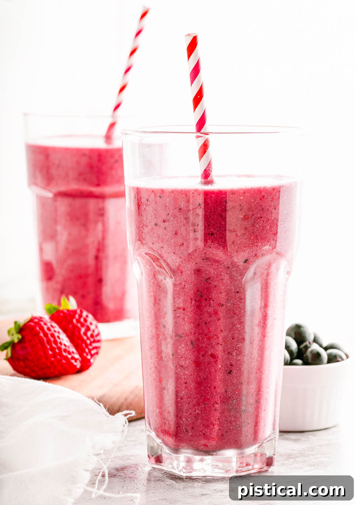 two glasses filled with berry smoothies sitting on and next to a small wood board. both smoothies have striped paper straws in them.