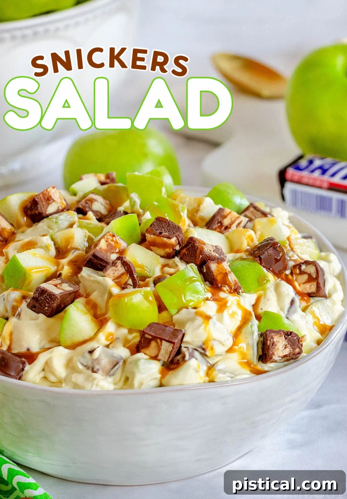 snickers salad in white salad bowl topped with a drizzle of caramel sauce. green apples and snickers candy bar in background. title overlay at top of image.