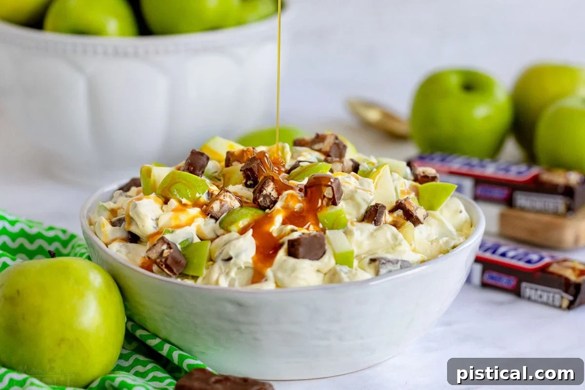 caramel sauce being drizzled over the top of the snickers salad in large white bowl. apples and snickers around the bowl.