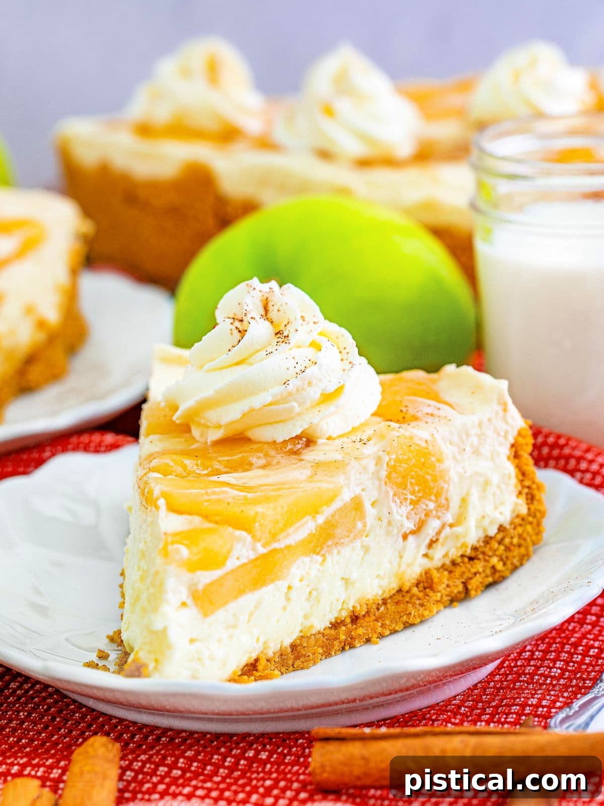 A slice of elegant no bake apple cheesecake on a white plate with a fork, garnished with whipped cream and a sprinkle of apple pie spice. The full cheesecake sits invitingly in the background.