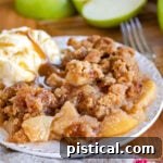 apple crumble served on a small white plate with vanilla ice cream and cut Granny Smith apples in background.
