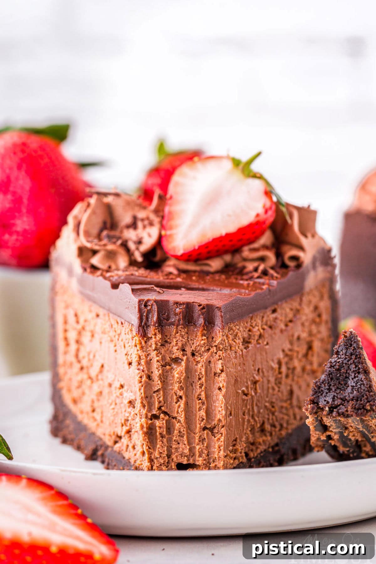 Slice of decadent chocolate cheesecake with an oreo crust and rich chocolate ganache on white round plate. Topped with chocolate shavings and fresh strawberries.