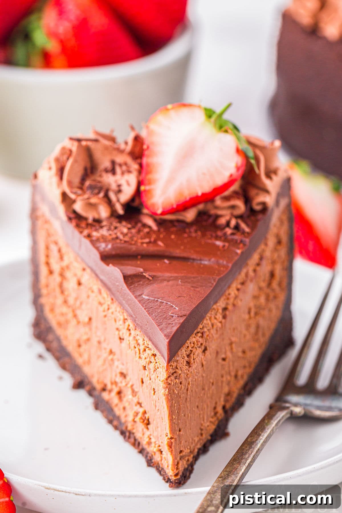 Slice of decadent chocolate cheesecake with an oreo crust and rich chocolate ganache on white round plate. Topped with chocolate shavings and fresh strawberries. Strawberries and more cheesecake can be seen in the background.