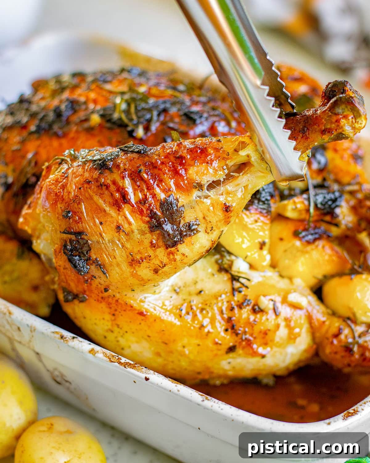 tongs pulling up on leg from a whole roasted chicken still in the roasting dish.