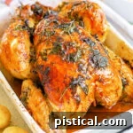 roasted chicken with golden skin and herbs cooked on top sitting in white roasting dish.