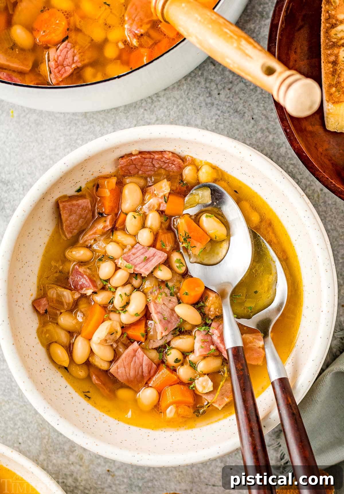 bowl of ham and bean soup with two spoons with wood handles next to pot of soup.