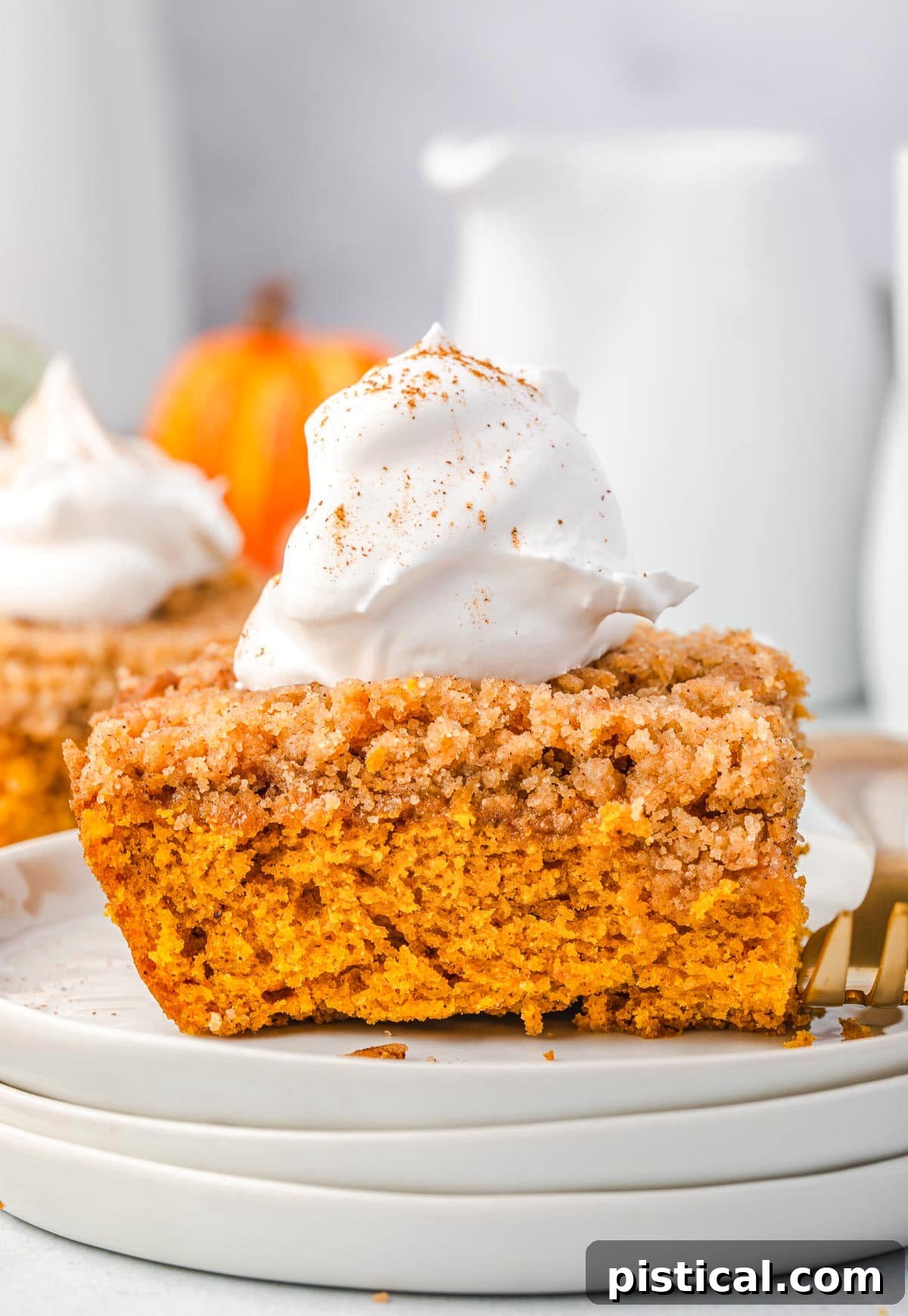 A generous slice of pumpkin coffee cake, elegantly placed on a stack of pristine white plates, is adorned with a dollop of fresh whipped cream, ready to be savored.