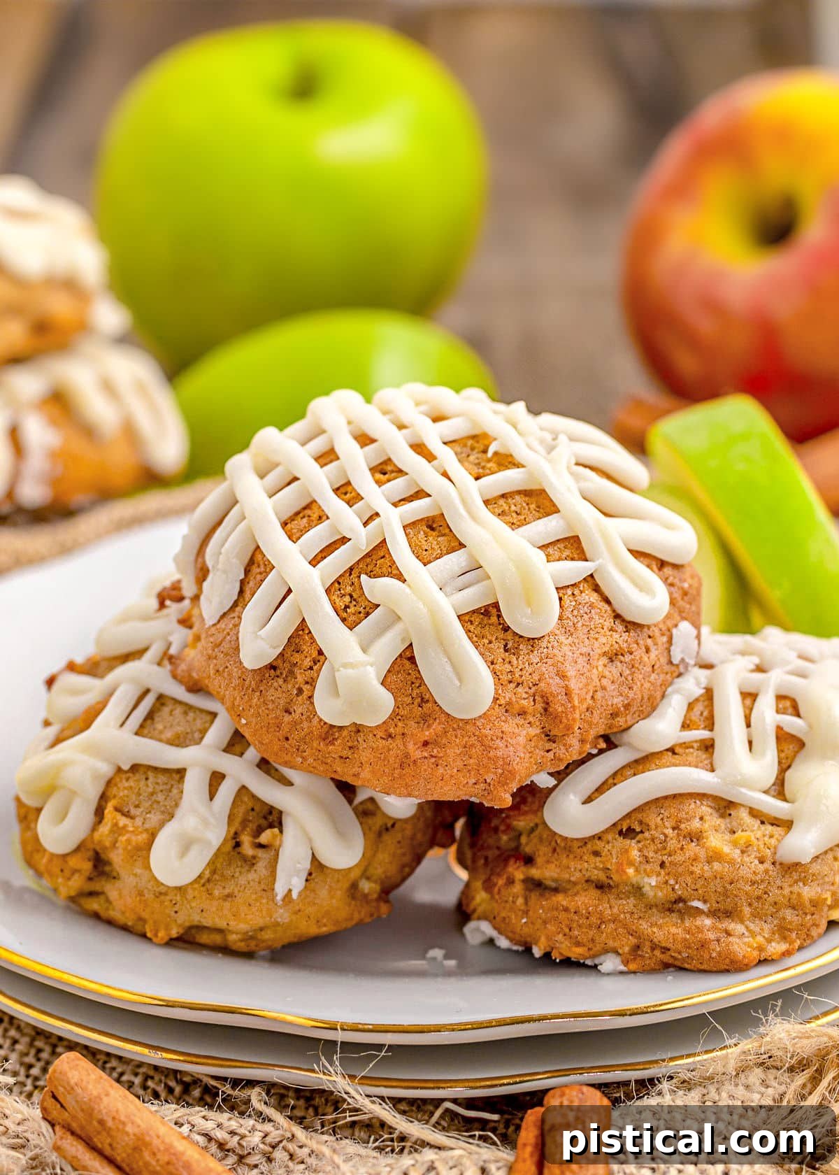 Three maple-iced apple cookies arranged on a white plate with a gold rim. Fresh apples and apple slices provide a rustic background, highlighting the seasonal theme.