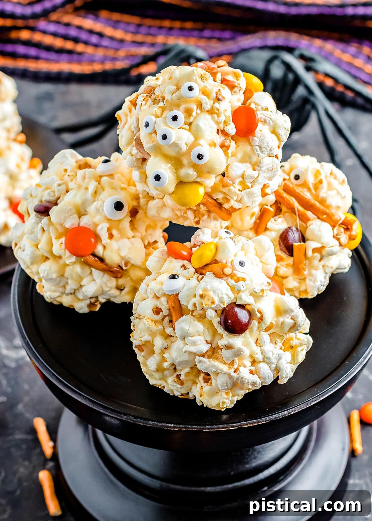 Assorted Halloween Popcorn Balls, whimsically decorated with candies and pretzel pieces, presented on an elegant black cake stand, ready for a festive celebration.