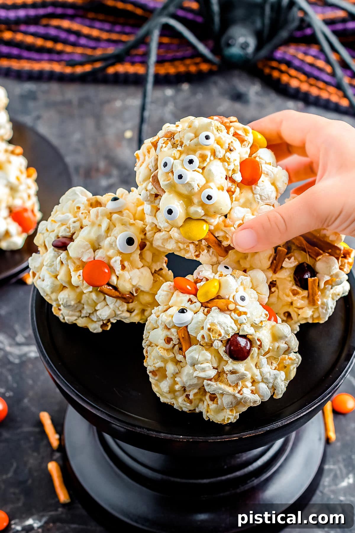 A hand gently reaching to pick up a single, artfully decorated Halloween popcorn ball from a tiered black cake stand, highlighting its festive appeal.