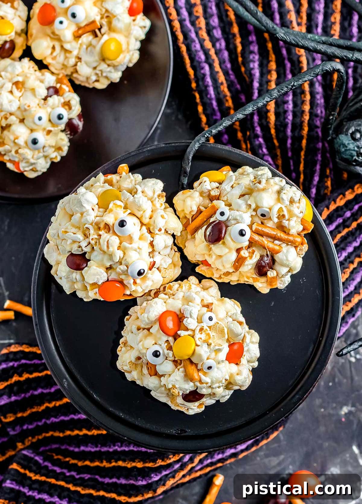A top-down perspective of three vibrantly decorated Halloween popcorn balls arranged on a dark cake stand, showcasing their festive candy embellishments.