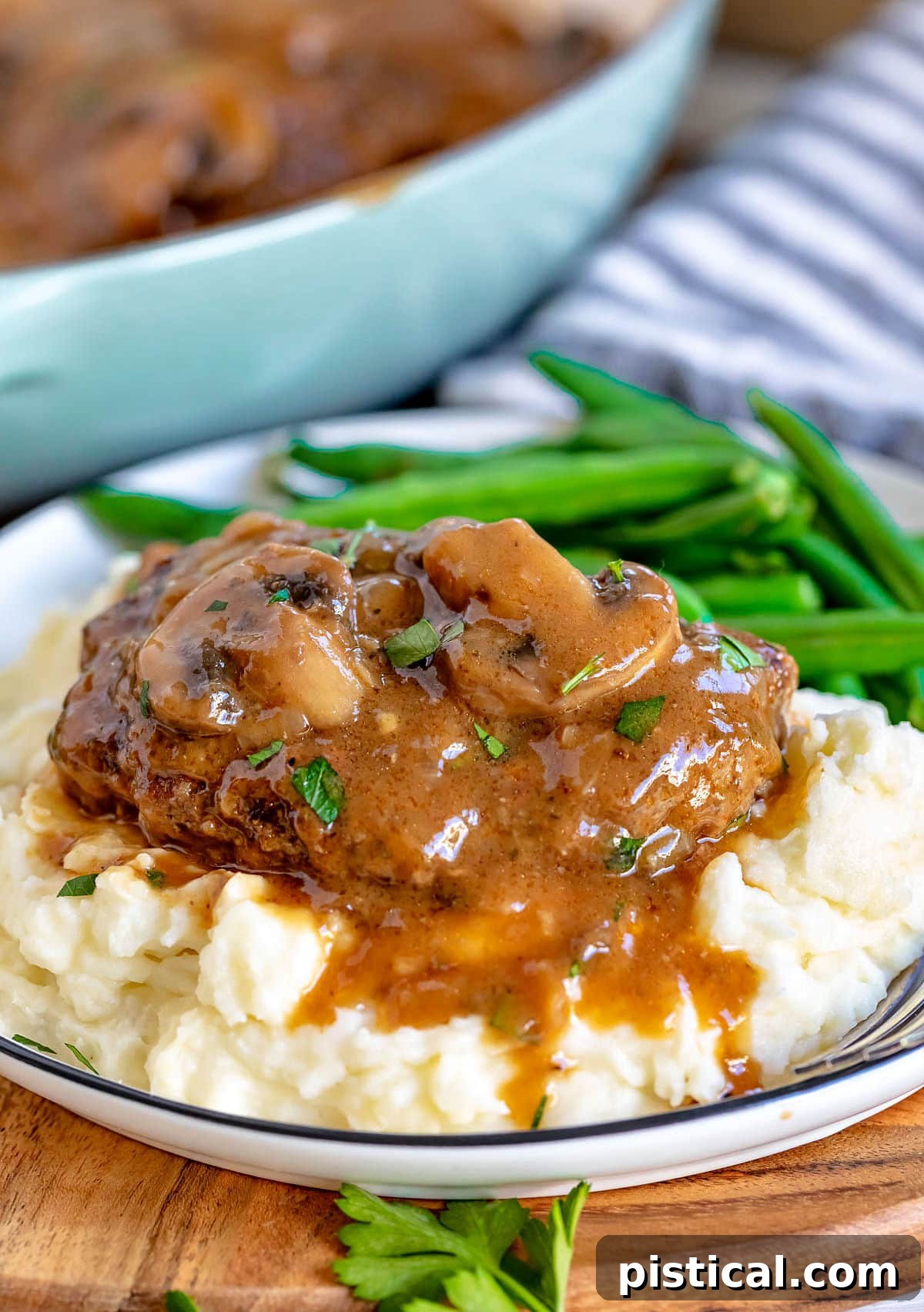salisbury steak on bed of mashed potatoes with green beans as well. Mushroom gravy is drizzled on top of the steak and mashed potatoes.