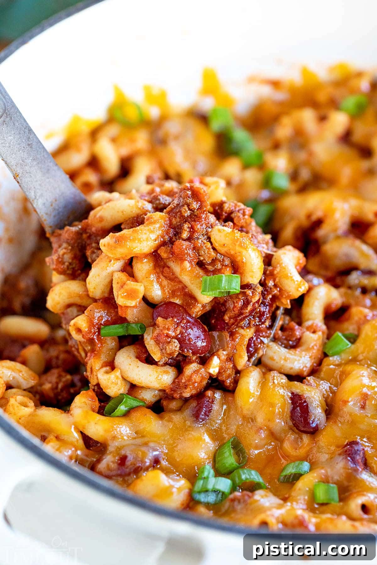 ladle of chili mac recipe in a white dutch oven topped with green onions.