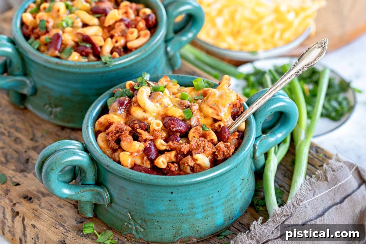 two bowls of chili mac topped with colby jack cheese and green onions. sitting on a thick wood board.