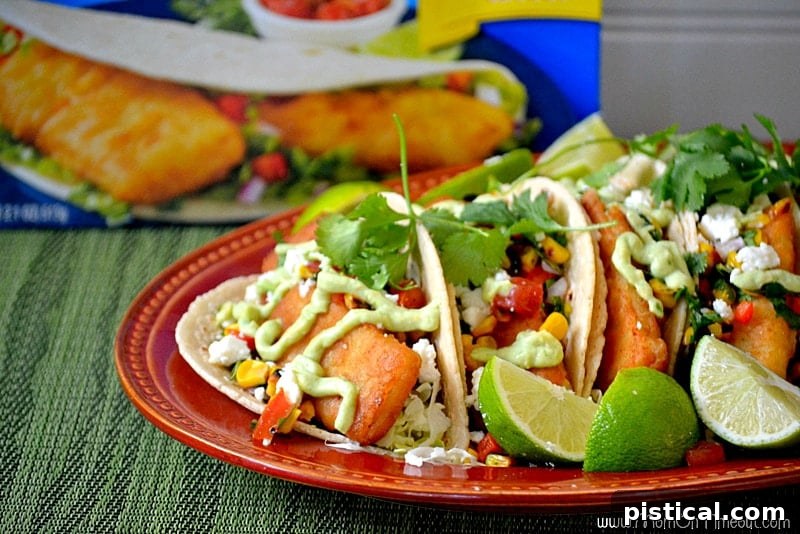 Overhead shot of two fish tacos on a platter with a side of roasted corn salsa and avocado sauce