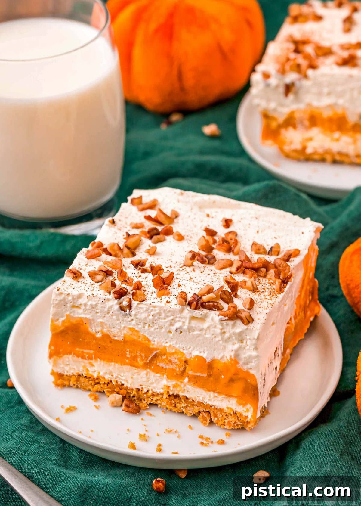 Top-down view of a four-layer pumpkin dessert on a white plate, garnished with toasted pecans and pumpkin pie spice. A glass of milk sits blurred in the background, suggesting a delightful pairing.