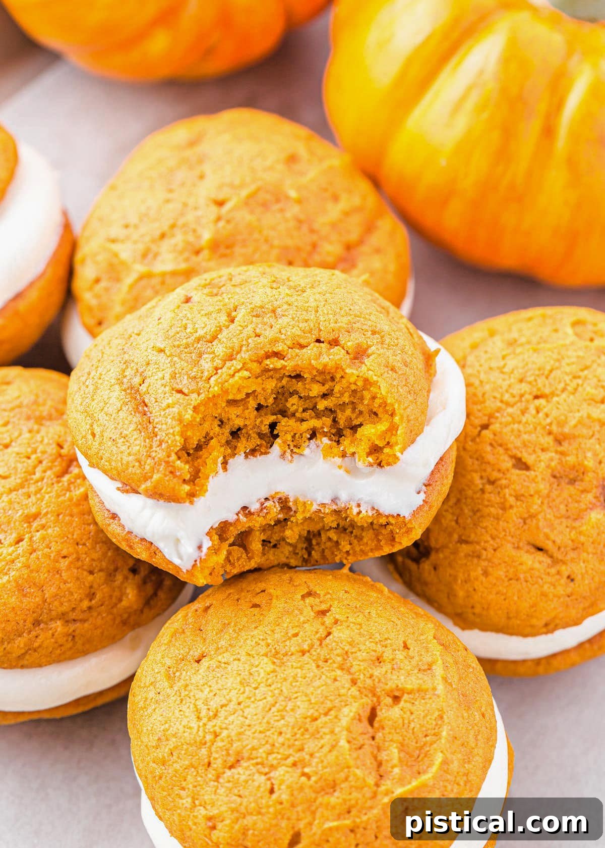 A close-up view of a pumpkin whoopie pie with a perfect bite taken out of it, revealing the thick, creamy filling. More whoopie pies are visible in the background, hinting at a bountiful batch of this delicious fall treat.