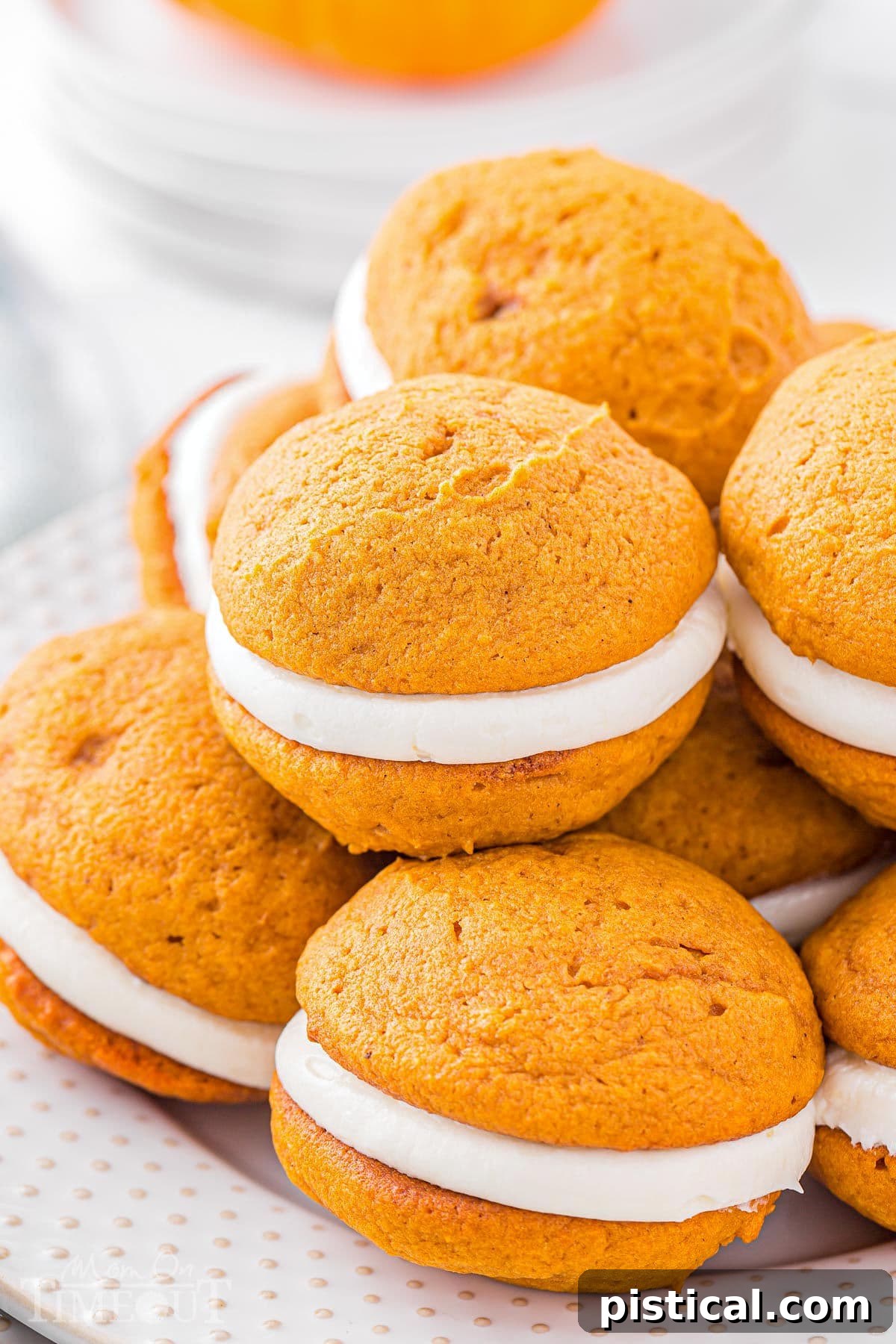 A dozen pumpkin whoopie pies are elegantly piled high on a pristine white plate, creating a tempting and abundant display. The soft, spiced pumpkin cookies are generously filled with a creamy white frosting, highlighting their irresistible appeal.