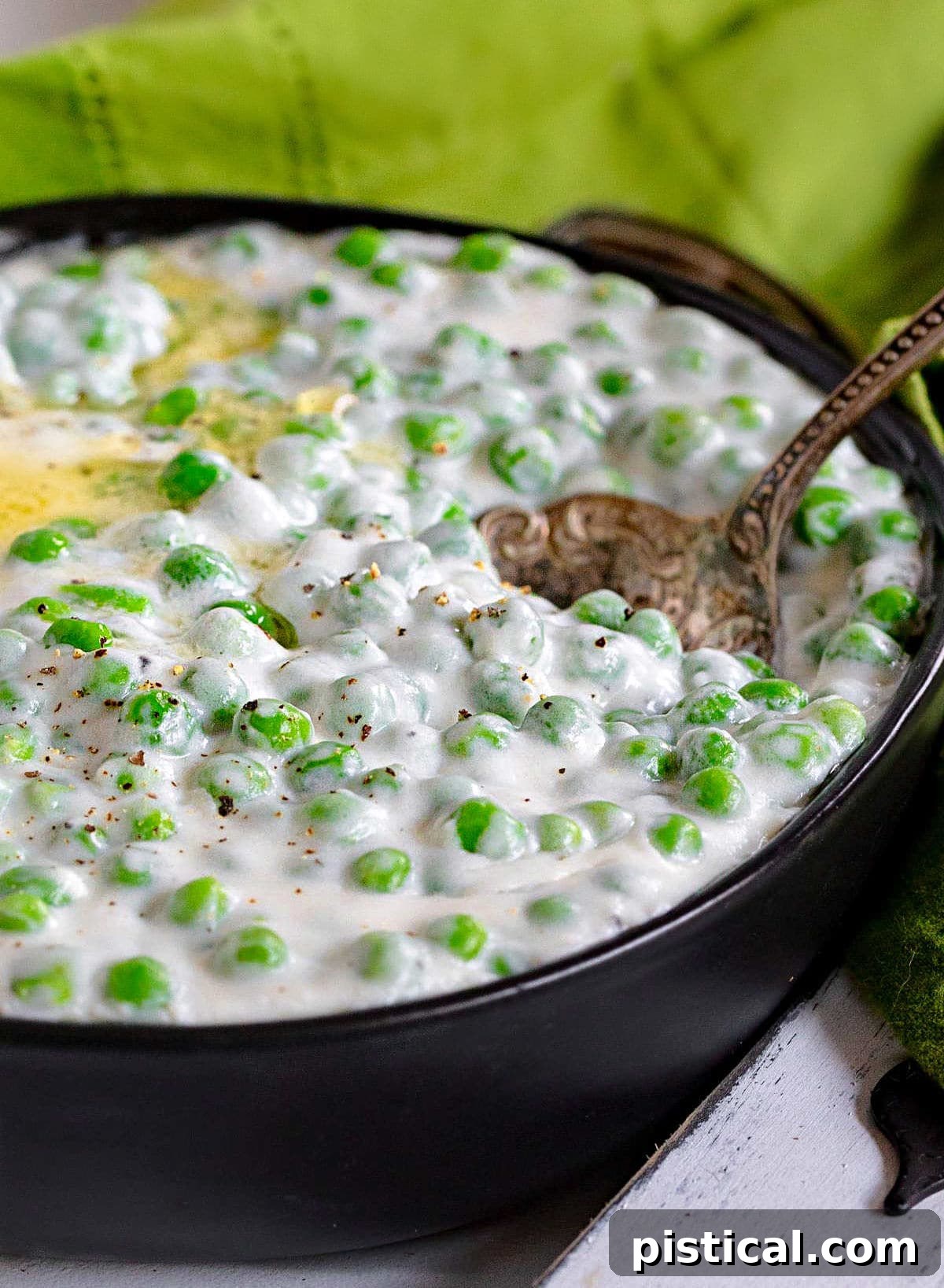 Creamed peas elegantly presented in a shallow black bowl, featuring a swirl of melted butter on top and an antique serving spoon resting within. A green napkin provides a soft background element.