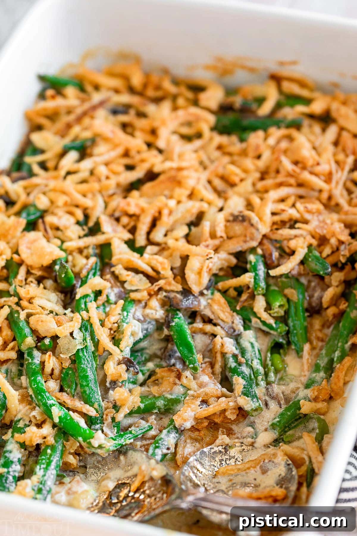 A large white baking dish filled with homemade green bean casserole, generously topped with golden-brown fried onions. Two silver serving spoons are nestled within the dish, suggesting a serving has just been taken, highlighting its irresistible appeal.