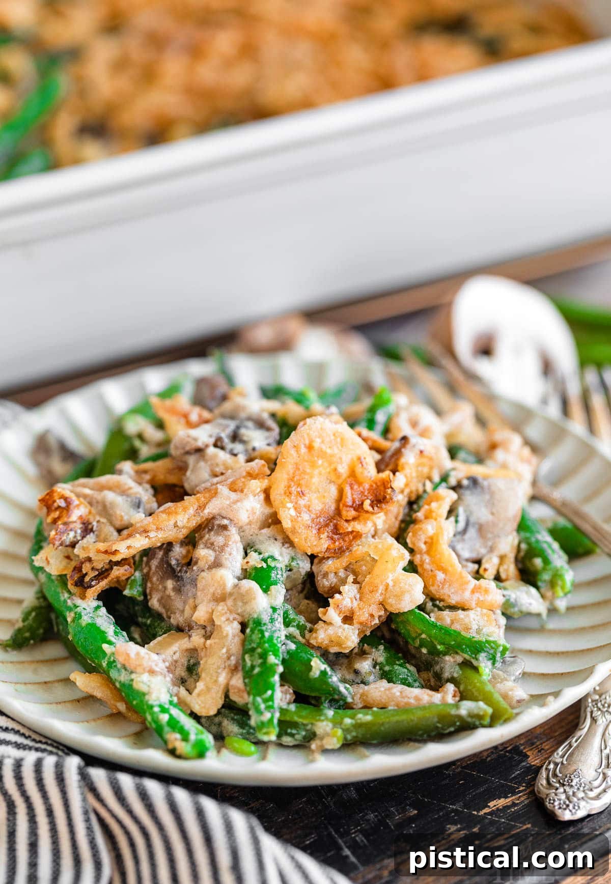 A small white plate holds a single serving of homemade green bean casserole, showcasing its creamy texture and crispy fried onion topping, with the larger casserole dish blurred in the background.