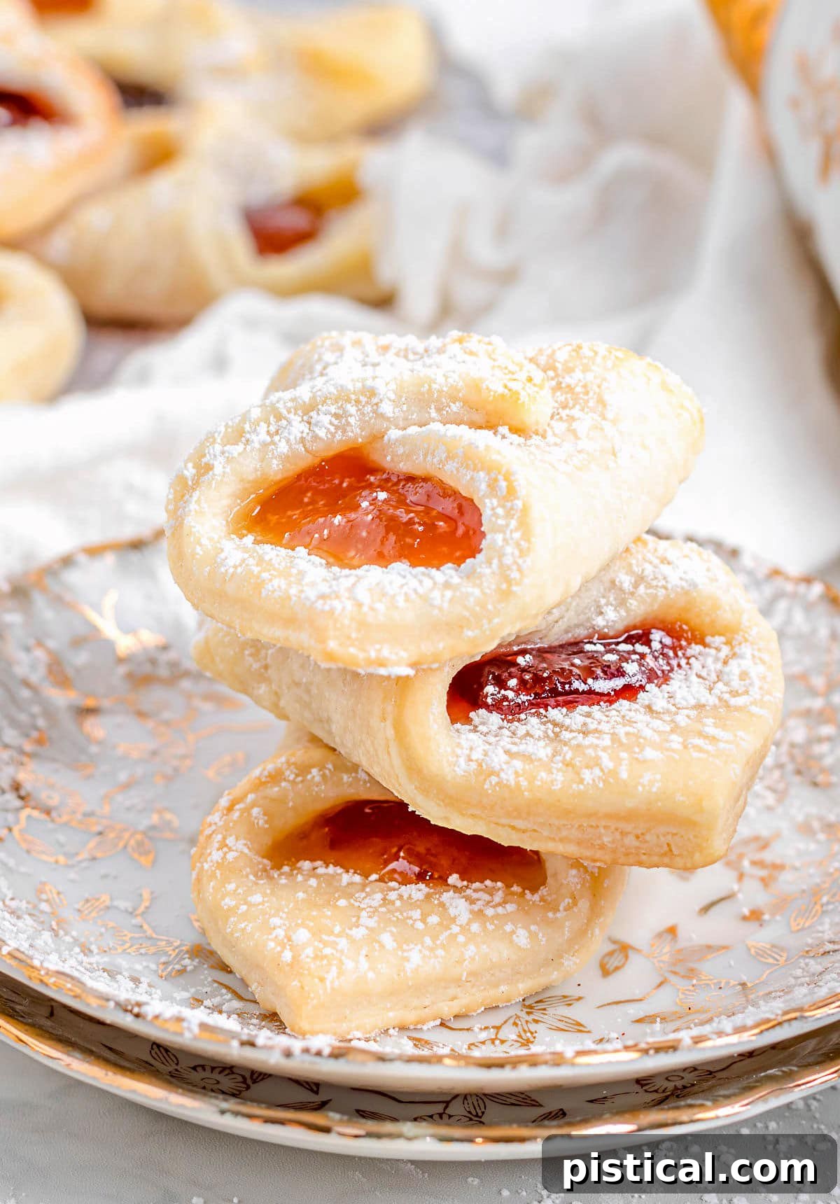 Kolaczki Beloved Polish Pastries 2 Three golden Kolaczki cookies, lightly dusted with powdered sugar, stacked on each other on two elegant gold-rimmed plates. One cookie reveals a sweet, tart jam filling.