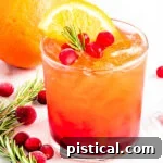 A stunning crystal glass filled with a vibrant red Christmas Punch, beautifully garnished with fresh rosemary, glistening cranberries, and a bright orange slice, set against a festive backdrop.
