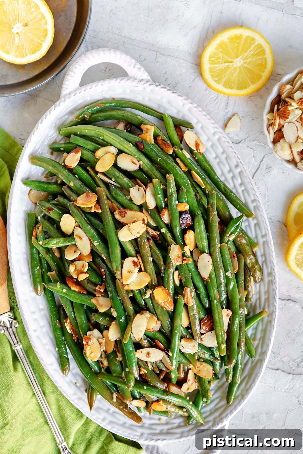 Top-down view of vibrant Green Beans Almondine generously topped with golden toasted almonds, served in a shallow white dish with fresh lemon slices artfully scattered to the side.