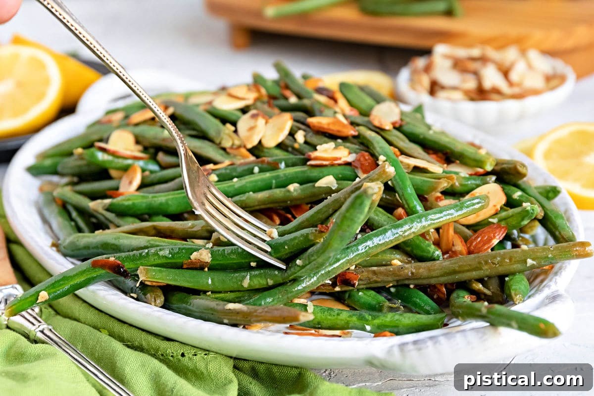 A forkful of Green Beans Almondine, rich with toasted almonds, poised above a shallow white serving dish. Fresh lemon slices and a neatly folded green cloth napkin are visible nearby.