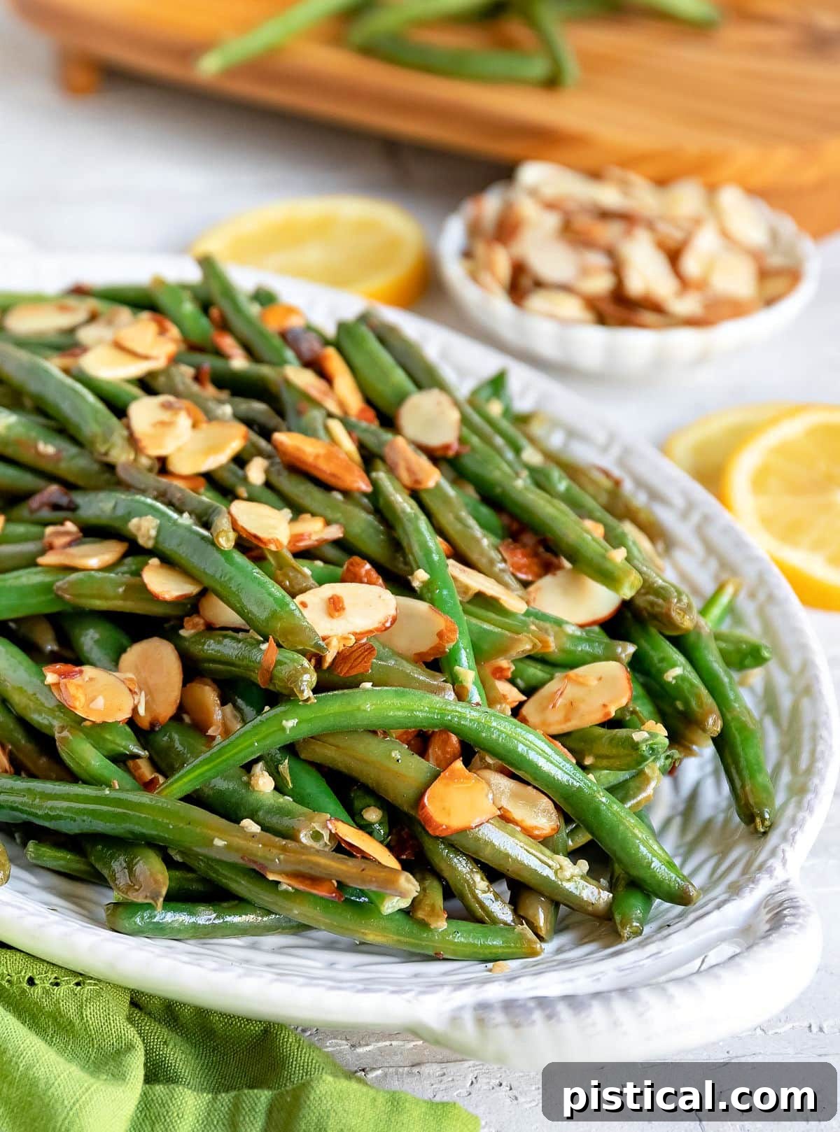 A generous portion of Green Beans Almondine, beautifully arranged in a large white serving dish and crowned with perfectly toasted almonds, ready to be served. A small bowl of extra almonds and two vibrant lemon slices are visible in the background.