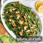 top down look at green beans almondine topped with toasted almonds on a shallow white serving dish with lemon slices scattered to the side.