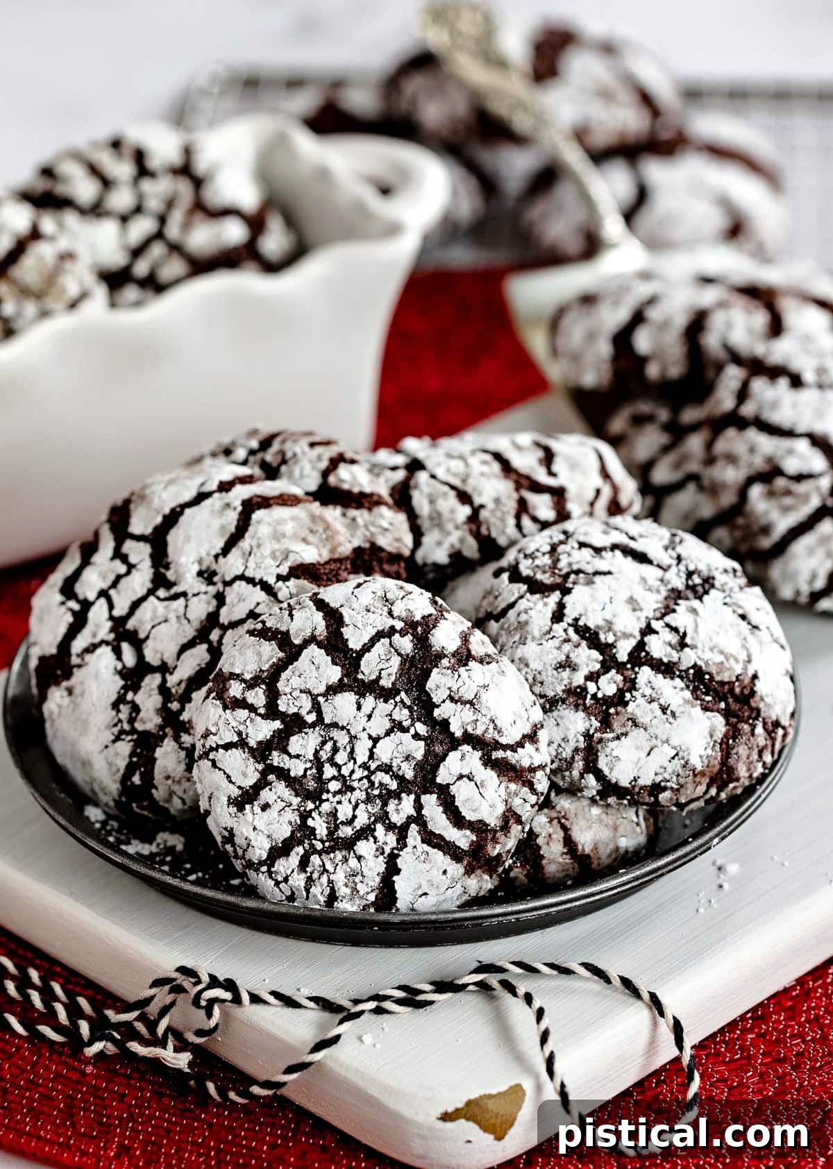 Small metal plate with six chocolate crinkle cookies sitting on a white wood board with more cookies on a server and cooling rack in background, illustrating their festive appeal.