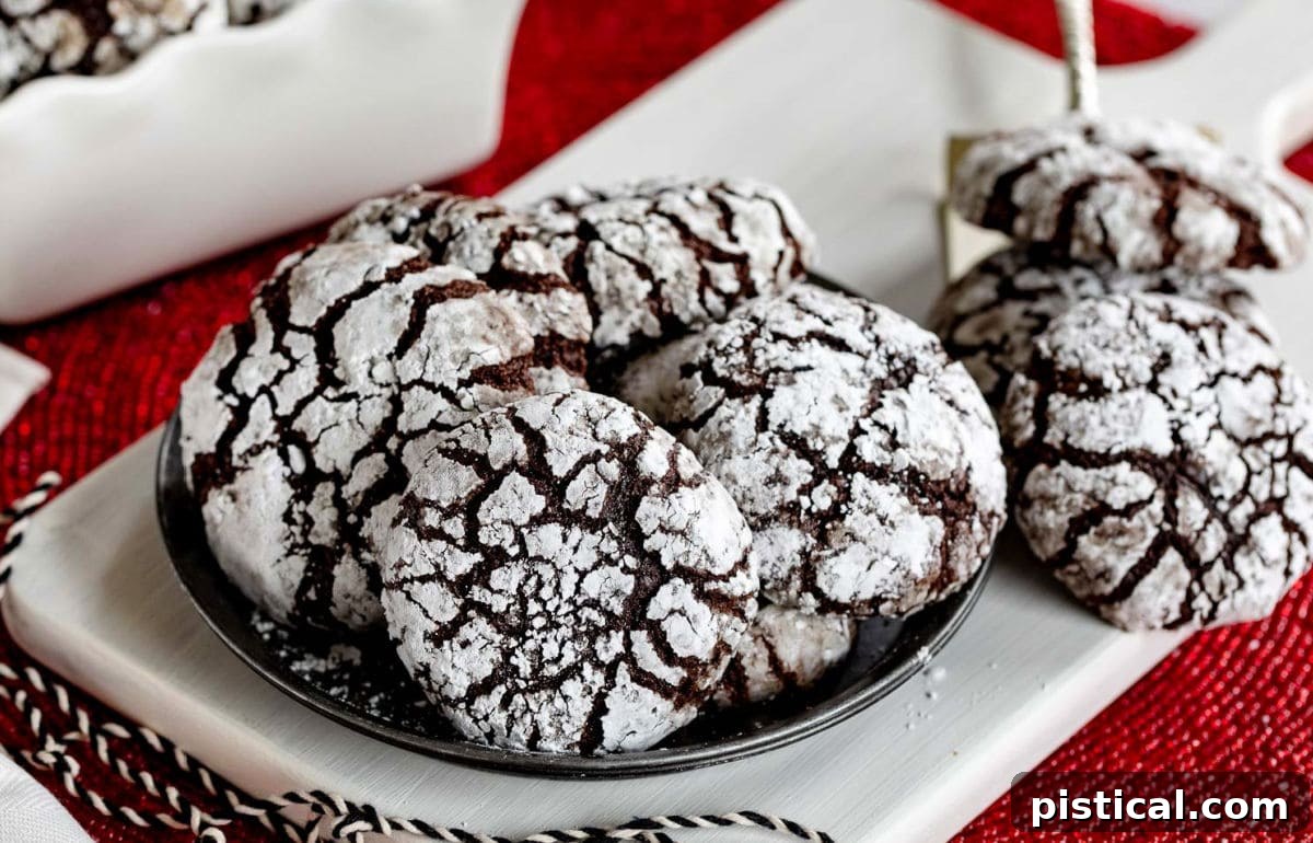 Plate of chocolate crinkle cookies with more cookies sitting off to the side and a few more in a white ceramic container, all on a red placemat. Perfect for holiday gatherings.