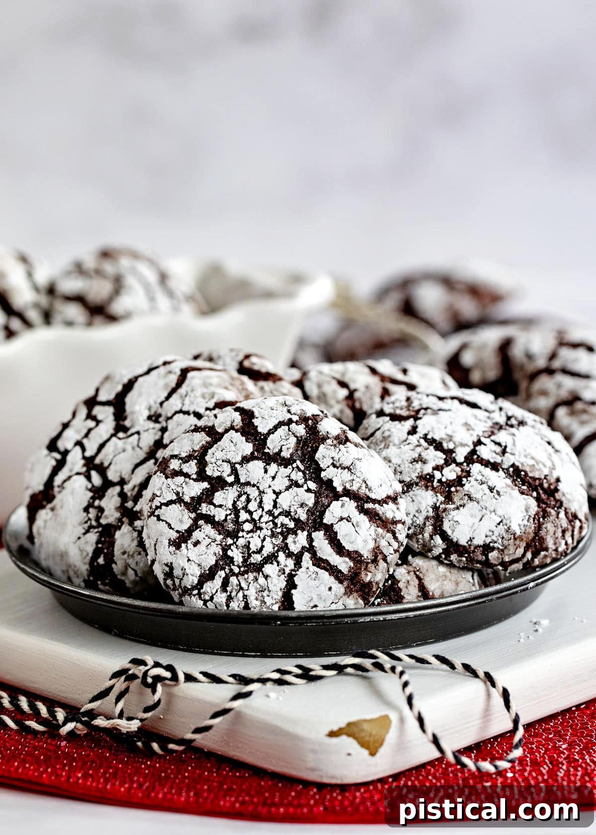 Plate of chocolate crinkle cookies sitting on white wood board on red sparkly placemat. Black and white twine sitting on board and more cookies can be seen in the background, creating a cozy holiday ambiance.