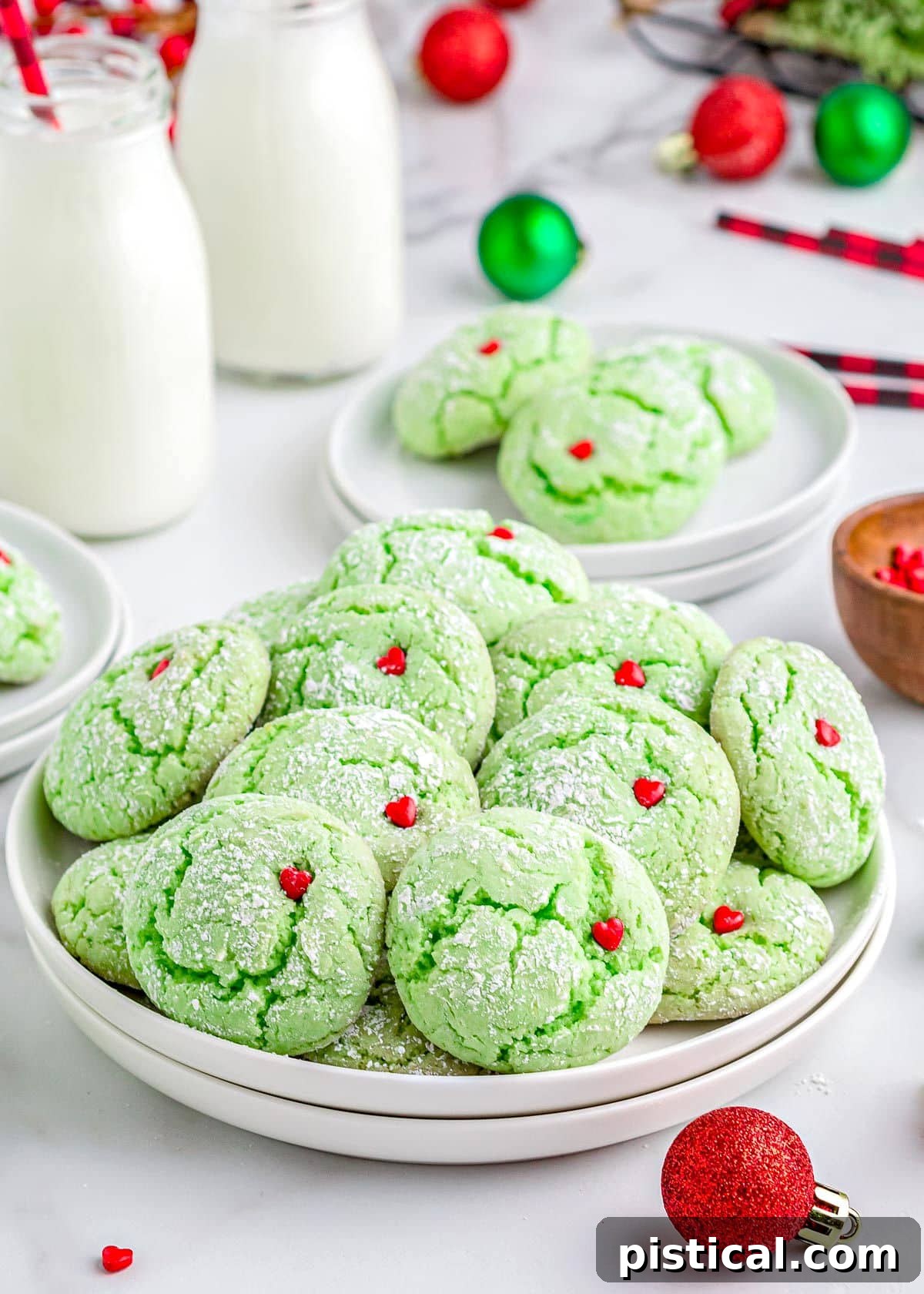 Vibrant green Grinch Cookies topped with bright red heart sprinkles, arranged on a white plate with milk jars and festive Christmas ornaments in the background.