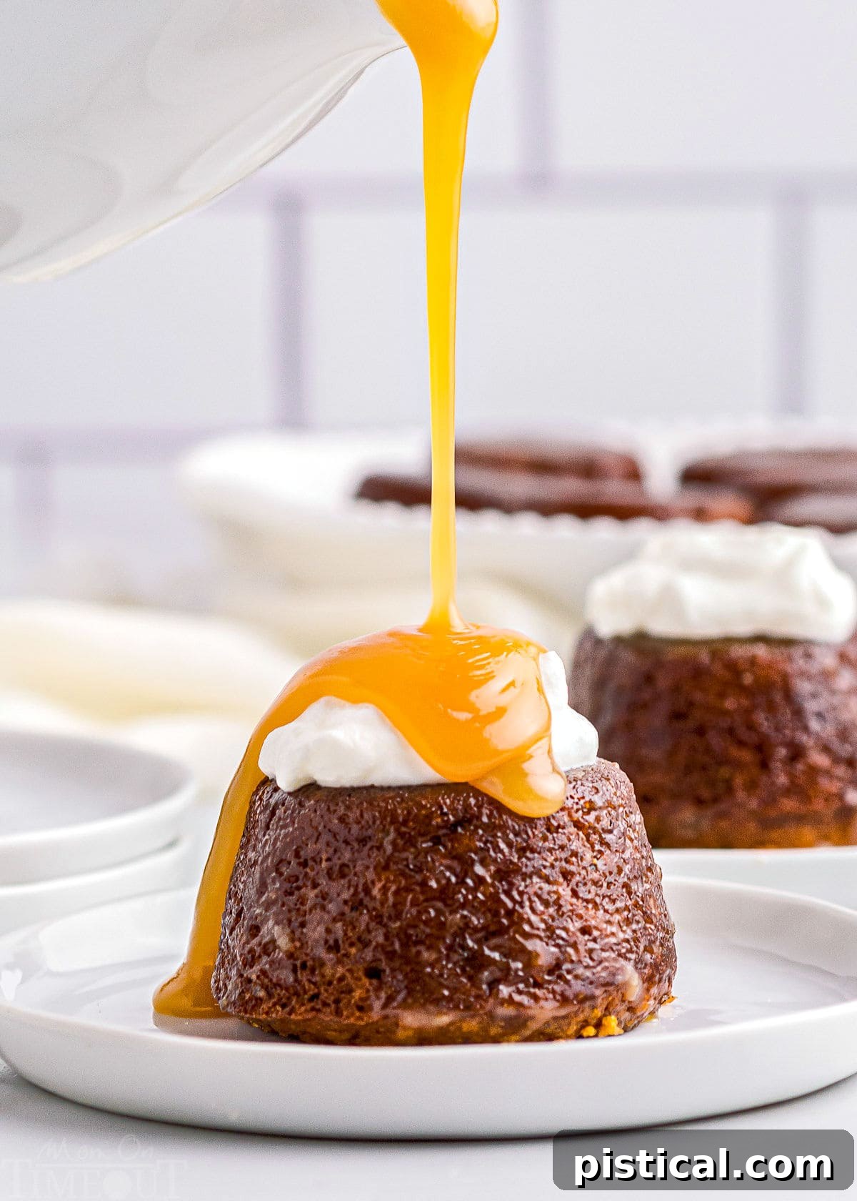Rich toffee sauce being generously poured over a sticky toffee pudding, already adorned with whipped cream, on a white dessert plate.